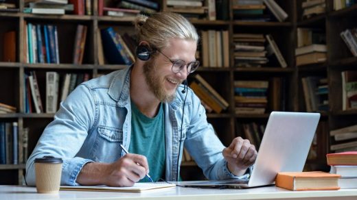 Smiling happy young man wear wireless headset look at laptop screen make notes study e learning in library watch webinar training online course video call, distance education, skype teaching concept