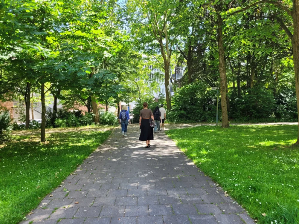 Hörsalon. Bild von City Soundspaces. Weg mit grünem Rasen links und recht. Grüne Bäume säumen den Rasen. Drei Menschen laufen mit dem Rücken zur Kamera von der Kamera weg.